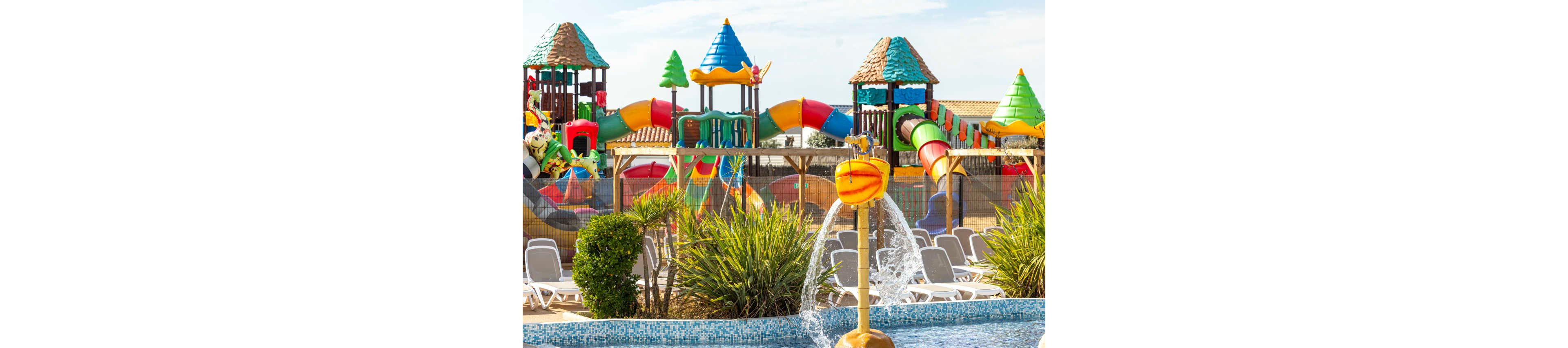 A vibrant outdoor water playground features colorful slides, tunnels, and climbing structures. In the foreground, a small pool with a playful water fountain splashes into a tiled basin. Sun loungers are arranged around the pool area, suggesting a relaxing family-friendly environment. Bright colors and clear skies create a cheerful, sunny holiday atmosphere.