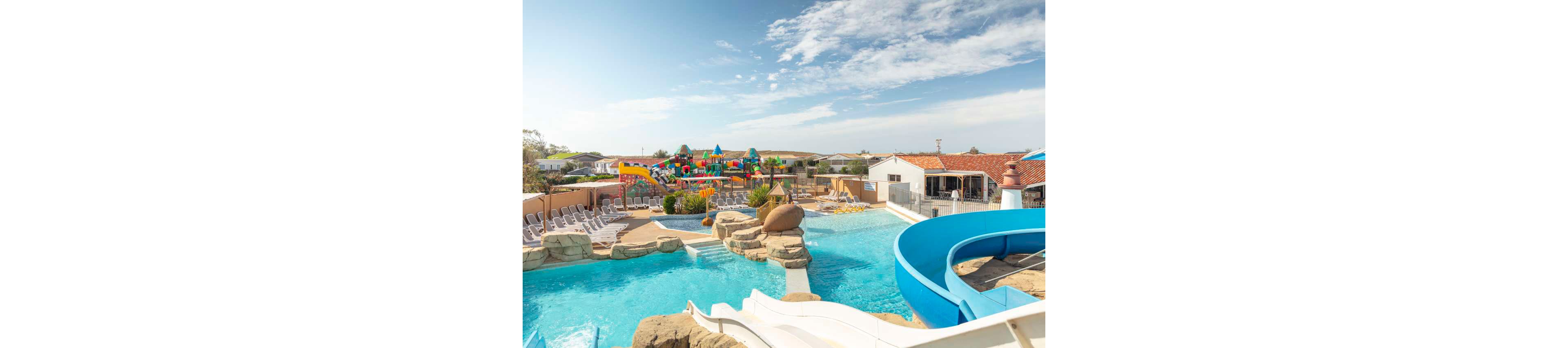 Large outdoor swimming pool complex featuring a curving blue waterslide and white slide in the foreground. Lounge chairs line the poolside deck beside artificial rock formations and shallow water areas. In the background, a colorful water playground with slides and climbing structures adds a family-friendly attraction. Low buildings with red-tiled roofs and open sky suggest a sunny vacation resort setting.