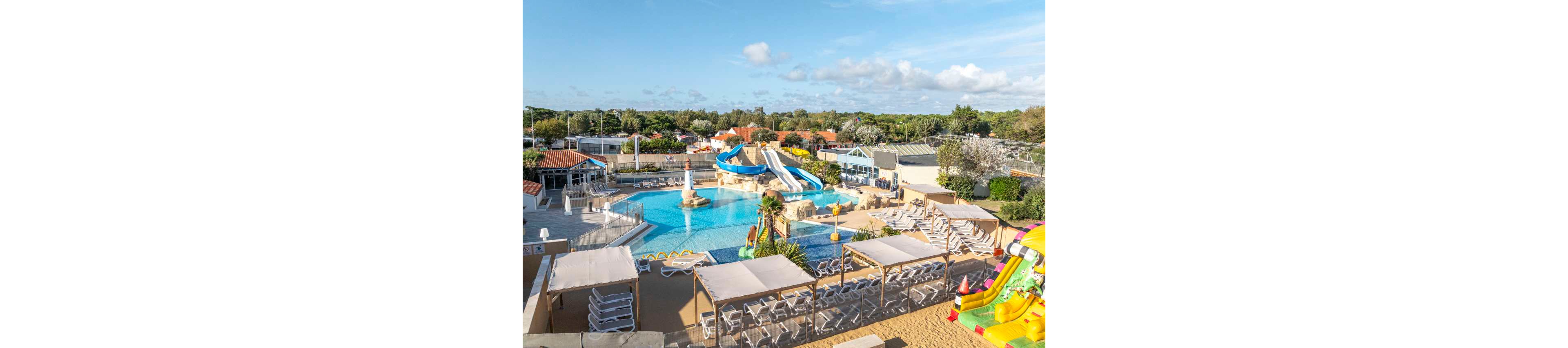 An outdoor water park features a large swimming pool surrounded by sun loungers and shaded cabanas. Several winding water slides in blue and white colors lead into the main pool area. A colorful inflatable slide structure is visible on the right side, adding to the family-friendly atmosphere. The scene is set on a bright, sunny day with blue skies and scattered clouds, and green trees in the background.