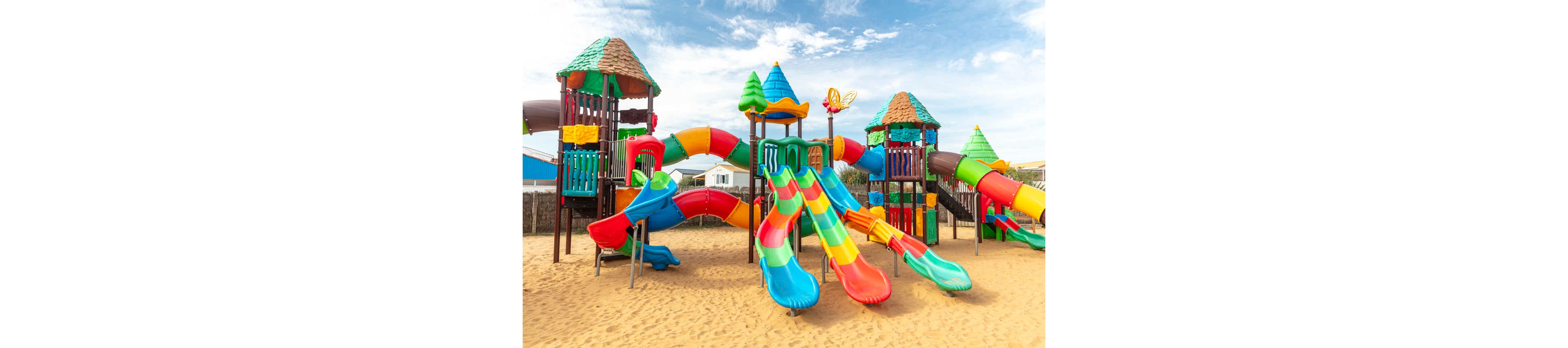 A large, colorful playground structure stands on a sandy surface under a bright blue sky. Multiple slides, tunnels, and climbing towers are connected in a playful maze. The equipment features vibrant red, yellow, green, and blue elements that create a cheerful atmosphere. No children are present, highlighting the design and layout of the play area.