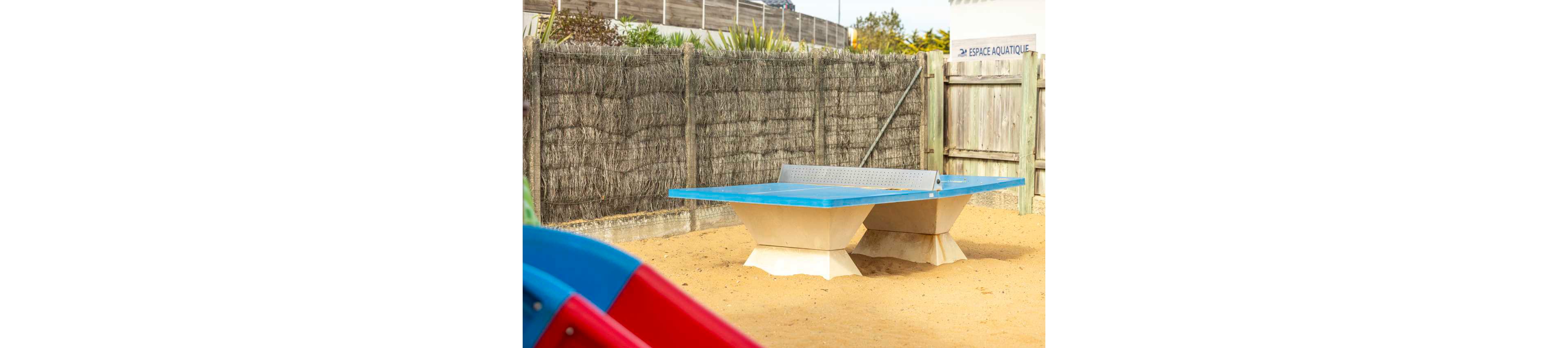 A sturdy outdoor ping pong table stands on a sandy surface in a fenced recreational area. The table has a blue top with a metal net and solid geometric concrete legs. In the background, tall woven fencing and wooden panels provide privacy, with some greenery visible above. A sign reading "ESPACE AQUATIQUE" suggests the area is near an aquatic or pool facility. Bright daylight highlights the clean, playful atmosphere of the space.