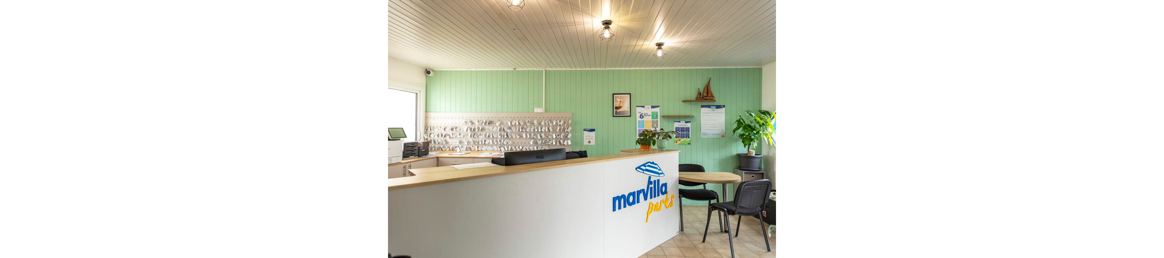 A clean, modern reception desk stands in a holiday park office with the name "marvilla parks" clearly visible on the front. Behind the counter, a large wall-mounted board displays numerous hanging keys, suggesting guest accommodations. The room features mint green wall paneling, light tiled flooring, and simple ceiling lights. Potted plants, wall posters, and a small model sailboat add decorative touches to the welcoming interior.