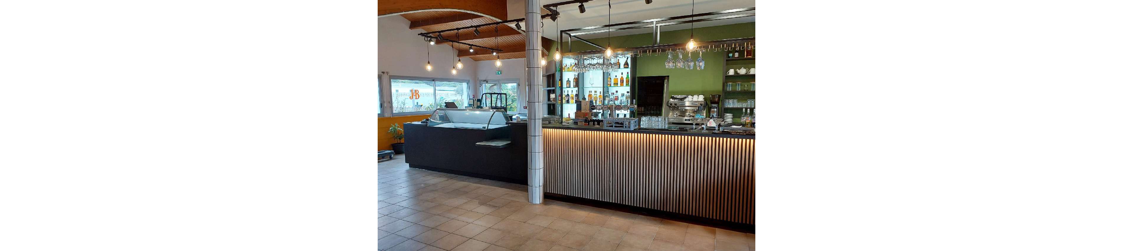Contemporary bar interior featuring a long illuminated counter with vertical slats and hanging pendant lights. Bottles and glassware are neatly arranged on shelves behind the bar, with a coffee machine and other equipment visible. An ice cream or dessert display case stands to the side near large windows that let in natural light. The space appears clean, quiet, and ready for customers, with tiled flooring and a mix of wood and metal finishes.