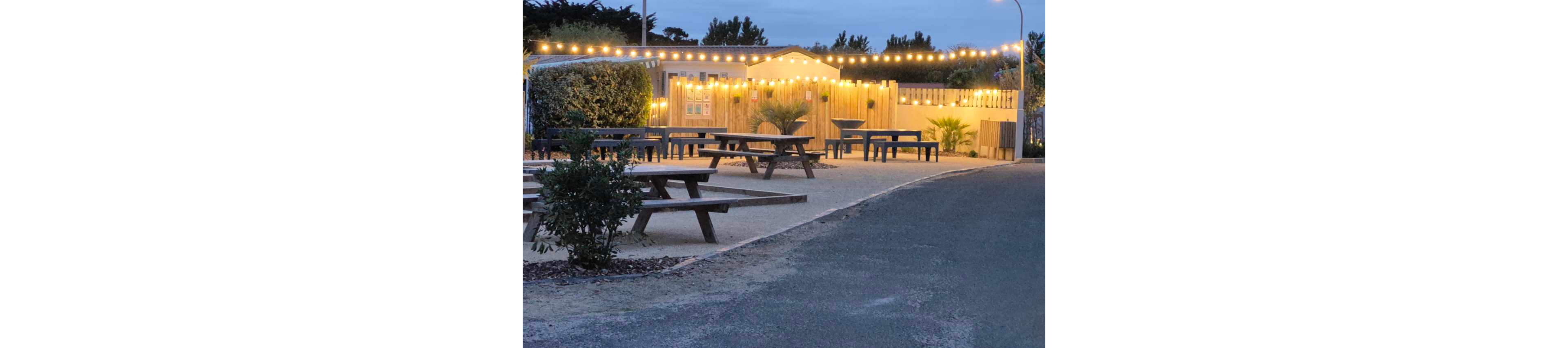 An outdoor seating area features several wooden picnic tables arranged on a gravel patio. Warm string lights hang above and around a small building, creating a cozy atmosphere at dusk. Potted plants and shrubs add greenery along the edges of the space. The scene is empty of people, with a quiet road curving past the illuminated terrace.