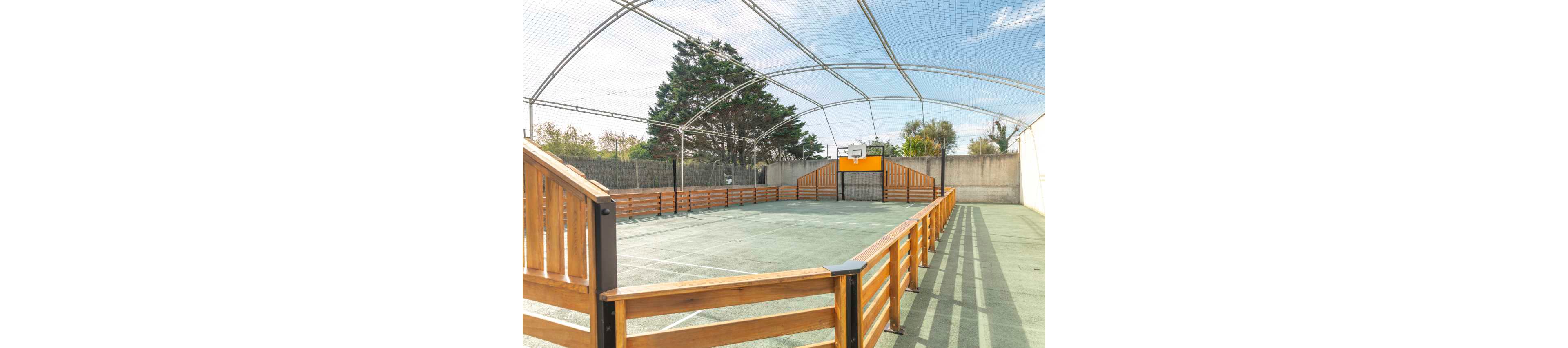 An empty outdoor multi-sport court is enclosed by wooden railings and high netting. A small basketball hoop and backboard are mounted at one end of the court. The playing surface appears to be a synthetic or hard court material under a clear sky. Trees and a tall concrete wall surround the sports area, creating a private recreational space.