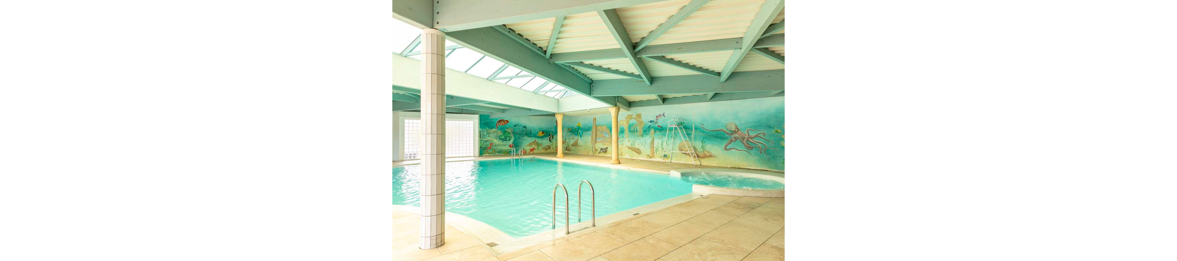 Indoor swimming pool with clear turquoise water is shown in a spacious, light-filled room. The walls feature colorful underwater murals with fish, seaweed, and ocean scenes. A metal ladder leads into the pool, and a small slide and separate spa area are visible in the background. Large windows and skylights allow natural light to brighten the pale tiled floor and painted ceiling beams.
