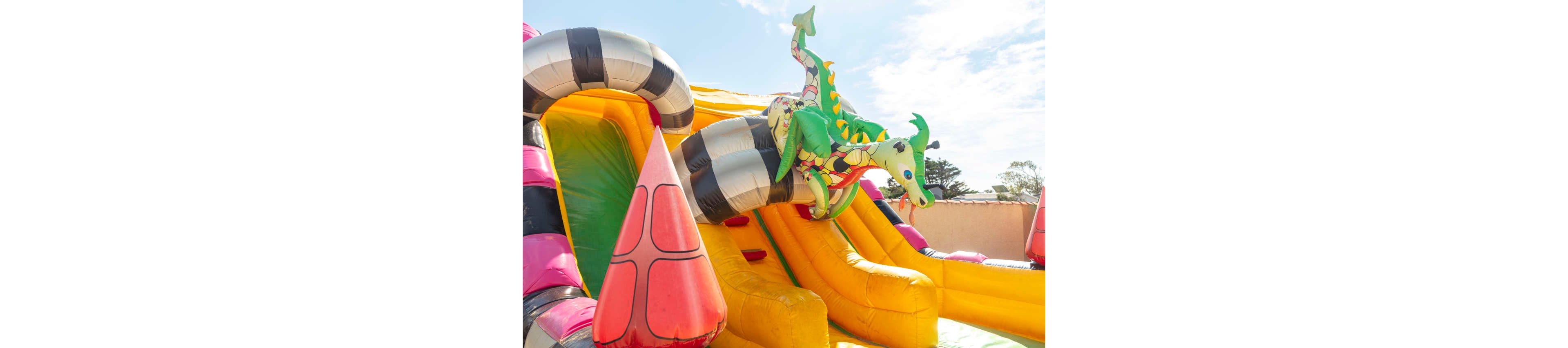 A brightly colored inflatable slide features a playful dragon figure wrapped around striped tubes. The structure is made of vivid yellow, pink, green, and black sections that stand out in the sunlight. It appears to be set outdoors on a clear day, with a blue sky and a few trees visible in the background. The scene suggests a fun, family-friendly attraction at a fair, festival, or amusement area.