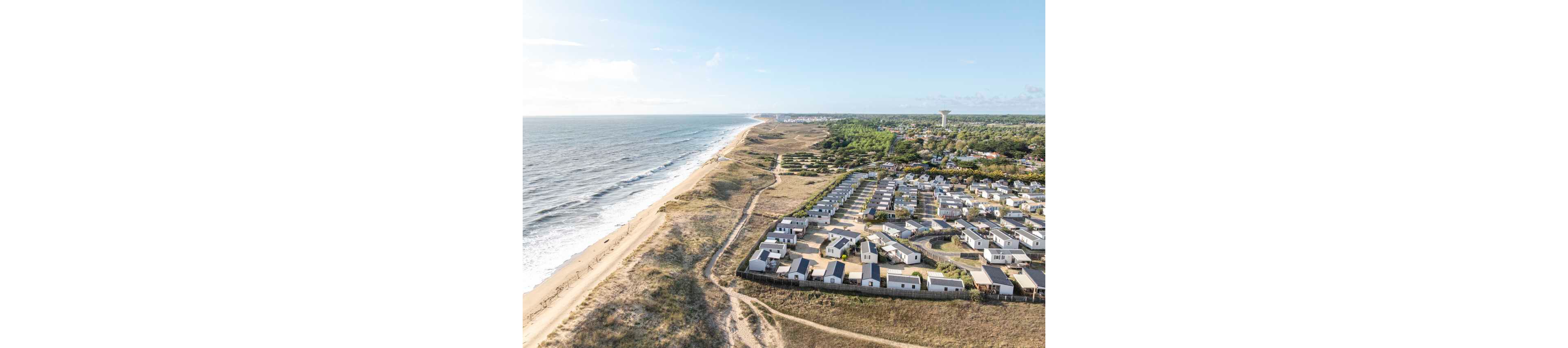 Aerial photograph of a coastal settlement with rows of small holiday homes near the shoreline. The cabins are arranged in a grid pattern beside sandy dunes and a long beach. Waves roll in from the ocean on the left, while green vegetation and a town extend into the distance on the right. The scene suggests a peaceful seaside vacation destination under a bright, partly cloudy sky.