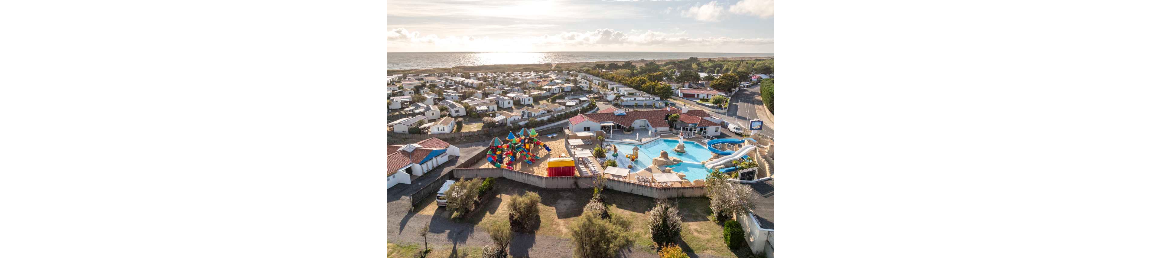 Aerial view of a coastal holiday park featuring numerous white mobile homes near the shoreline. In the foreground, a large outdoor swimming pool complex with slides and rock features is visible. A colorful playground with multiple shade structures stands beside the pool area. The scene is bathed in warm sunlight with the sea and horizon in the background, creating a relaxed vacation atmosphere.