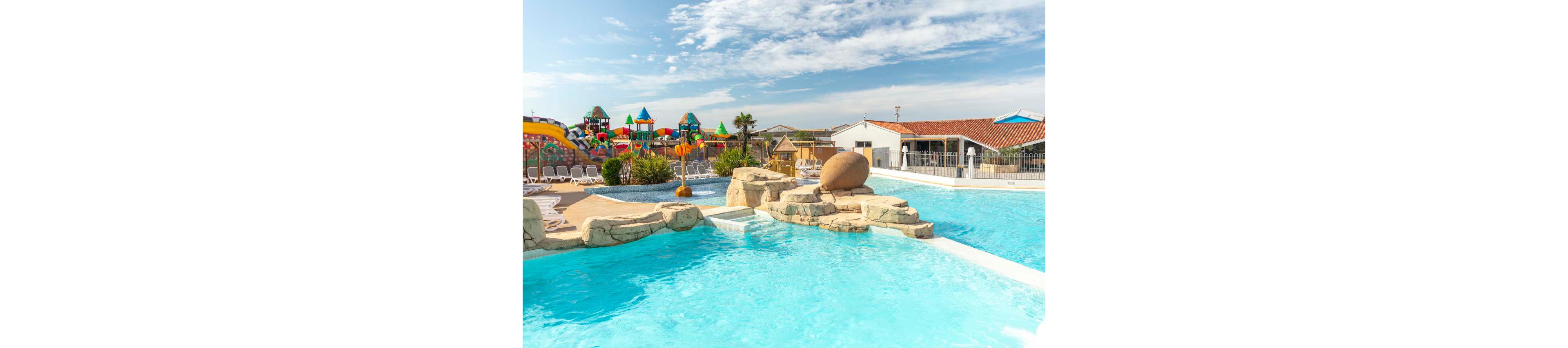 An outdoor swimming pool with clear blue water features rock formations and a shallow lounging area. In the background, a colorful water playground with slides and structures adds a fun, family-friendly atmosphere. Sun loungers line the poolside, and low buildings with red-tiled roofs suggest a holiday resort setting. The bright daylight and blue sky create a warm, inviting summer mood.