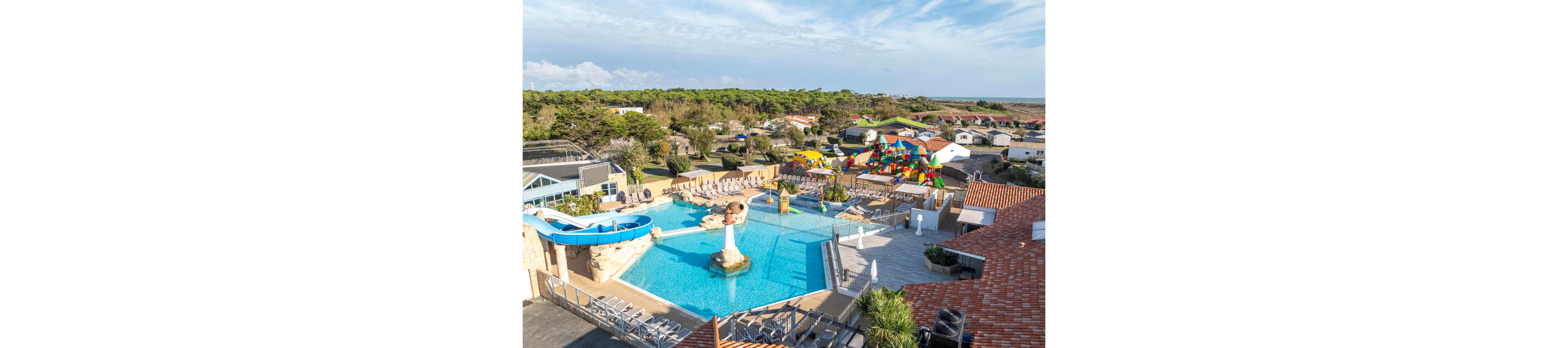 A large outdoor swimming pool complex is shown at a family holiday resort. The scene includes a blue waterslide, sun loungers, and a shallow pool area with playful water features. In the background, colorful inflatable play structures and cabins suggest a family-friendly campground setting. Trees, open sky, and distant coastline complete the relaxed vacation atmosphere.