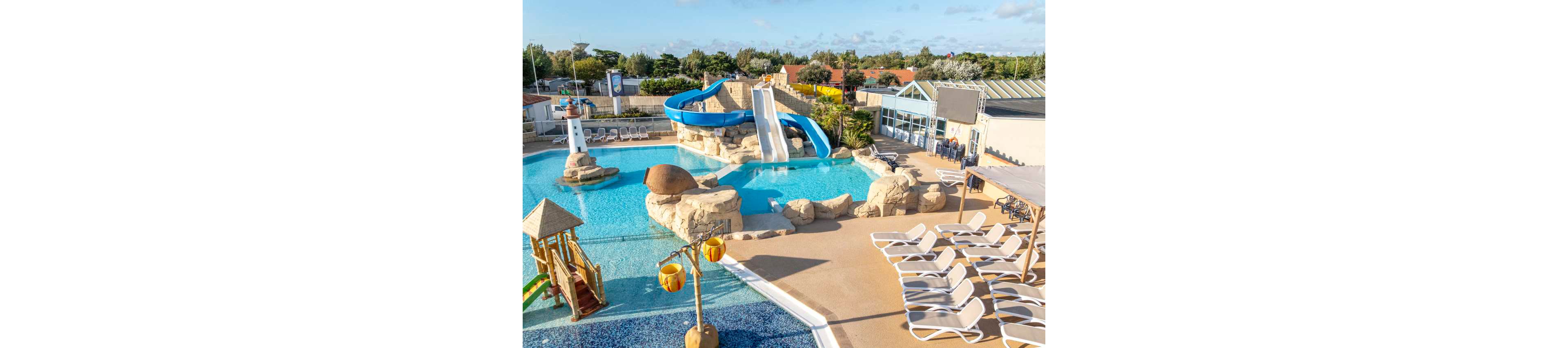 An outdoor resort-style swimming pool features large blue and white water slides curving into the clear water. Artificial rock formations surround the pool, creating a playful, lagoon-like setting. A small children's play area with a slide and splash zone sits in the foreground, while rows of empty sun loungers line the poolside deck. Buildings, trees, and a bright sky complete the relaxed holiday atmosphere.