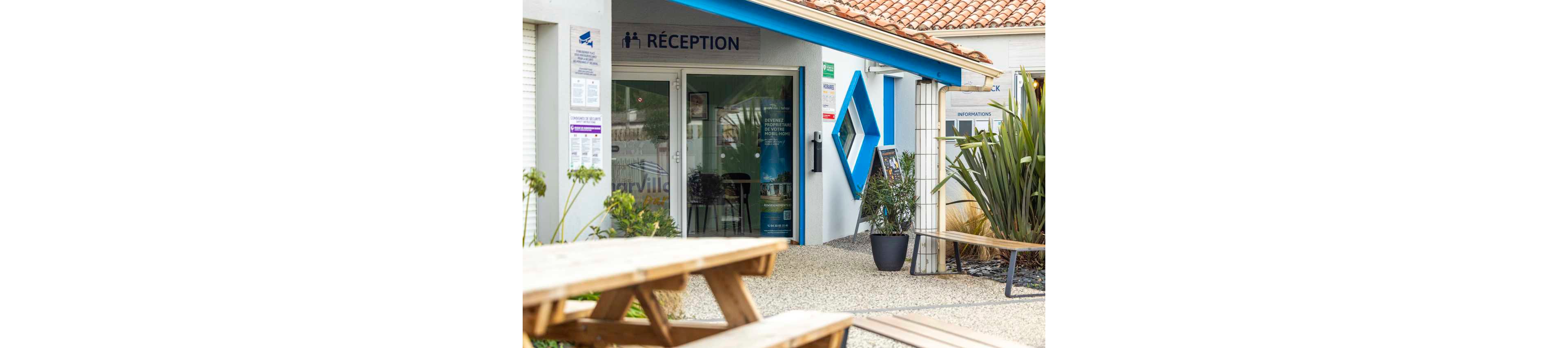 An outdoor reception building is shown with a glass entrance door and a sign reading “RÉCEPTION” above it. The setting appears to be a campsite or holiday park, with gravel ground and wooden picnic tables in the foreground. A large blue geometric wall feature and potted plants decorate the exterior, adding color and visual interest. Informational posters and signs are visible near the entrance, suggesting guest services and local information.