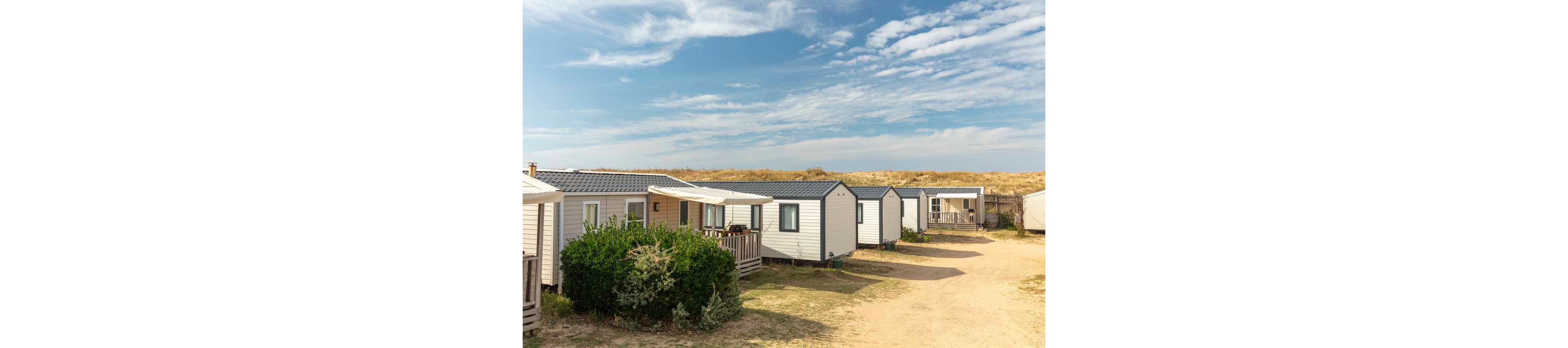 A row of light-colored mobile homes is arranged along a sandy path in a holiday park. The cabins feature small wooden decks and simple windows, with low shrubs planted in front of some units. In the background, grassy sand dunes rise under a bright blue sky with scattered clouds. The scene suggests a quiet, relaxed vacation setting near the coast.