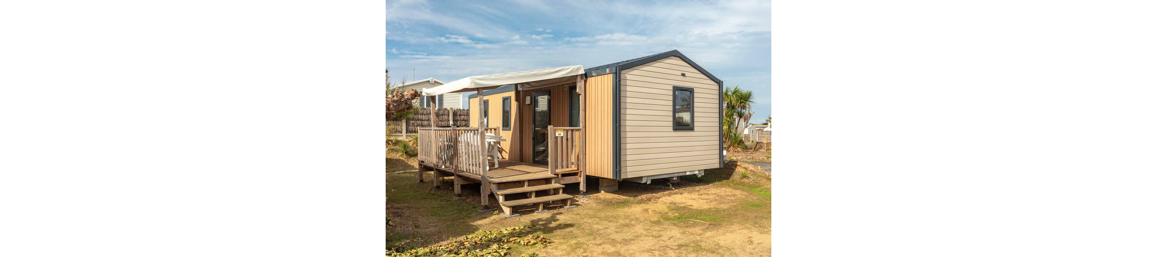 A modern mobile home stands on a dry, grassy plot with a small wooden deck and steps leading to the entrance. The structure features light-colored siding with contrasting wooden panels and a dark roof trim. An awning provides shade over the deck, where outdoor furniture is partially visible. Other similar units and a few palm-like plants can be seen in the background under a bright, partly cloudy sky.