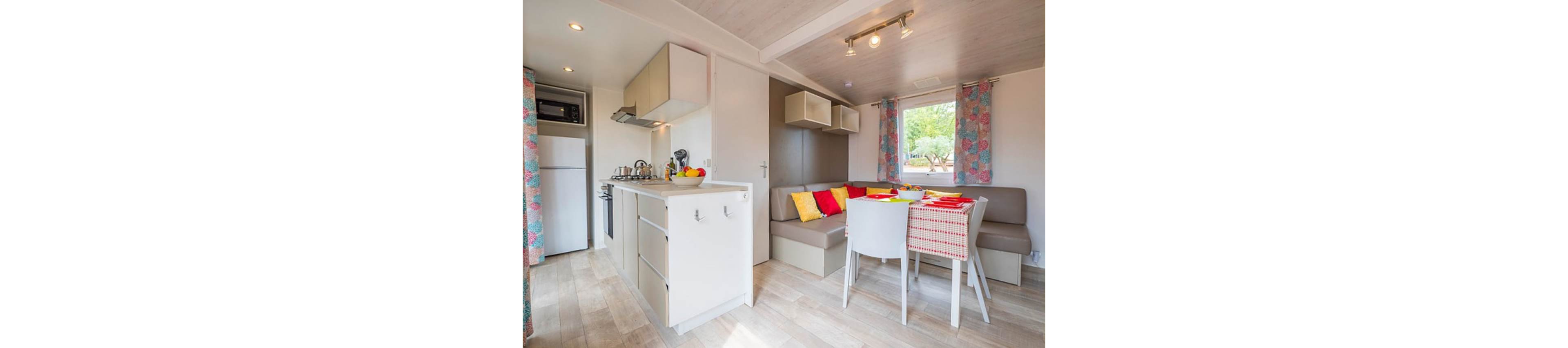 Cozy open-plan interior featuring a small kitchen and adjacent dining space. The kitchen includes white cabinets, a refrigerator, microwave, and stovetop with kettles and cookware. A corner sofa with colorful red, yellow, and patterned cushions sits beside a window with patterned curtains. A dining table with a red and white checkered tablecloth and white chairs completes the inviting holiday home atmosphere.