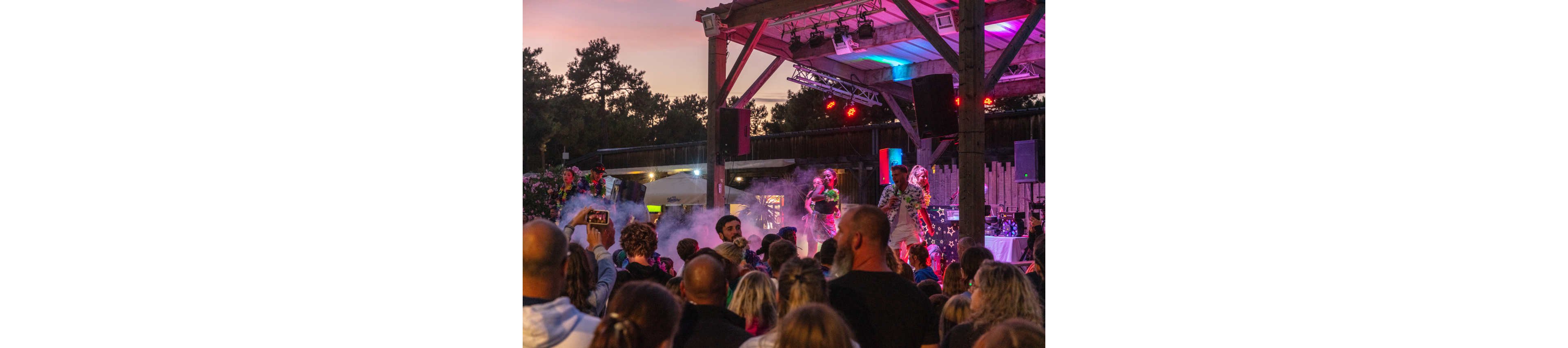 A large crowd gathers at an outdoor concert during dusk, watching performers on a covered stage. The scene is lit with colorful stage lights in pink, blue, and purple tones, creating a festive atmosphere. Smoke effects rise around the performers as people in the audience take photos and videos with their phones. Trees and wooden structures frame the venue, emphasizing the open-air setting and lively summer event vibe.