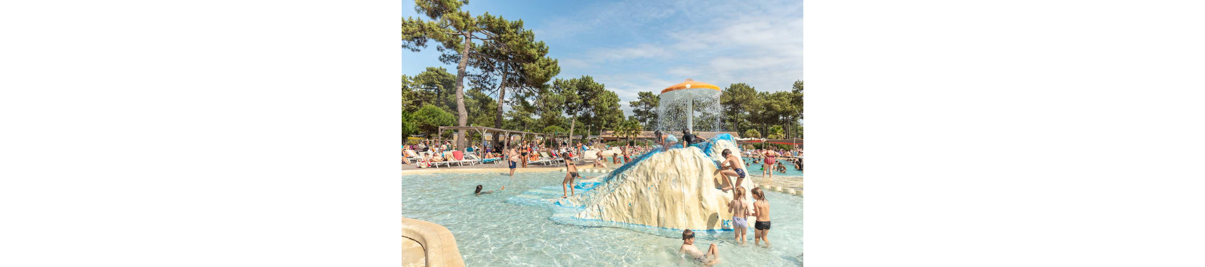 Children and adults play together in a shallow outdoor pool with a central climbing structure and waterfall feature. Several kids climb the artificial rock while others splash in the surrounding water. Sunbathers relax on loungers around the pool under clear skies. Tall pine trees frame the busy holiday scene, creating a relaxed summer vacation atmosphere.