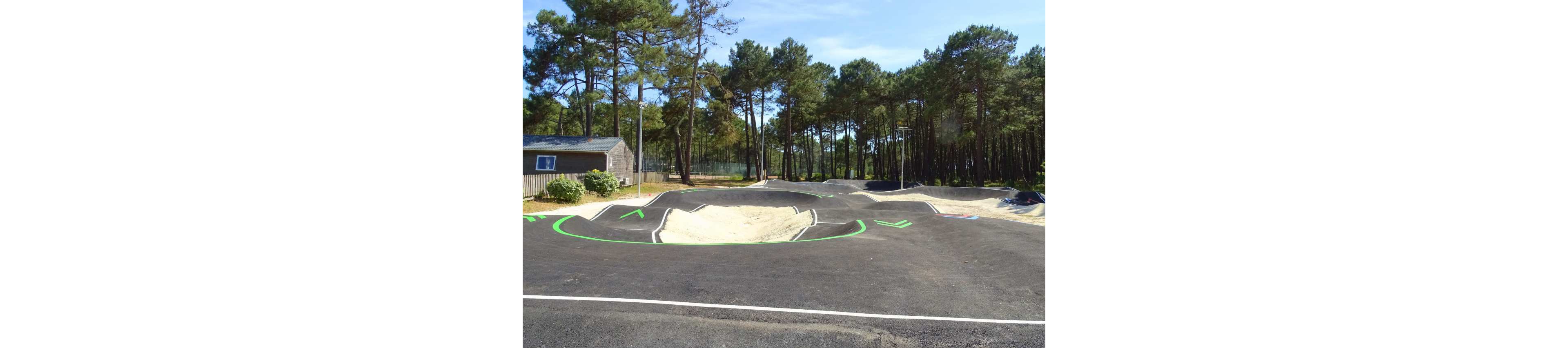 An empty asphalt pump track with multiple rolling hills and banked turns is set in a forested outdoor park. Bright green painted lines and arrows mark the riding path around the track. Sandy sections fill the center of some features, contrasting with the dark asphalt. Tall pine trees surround the area, and a small wooden building stands nearby under a clear sky.