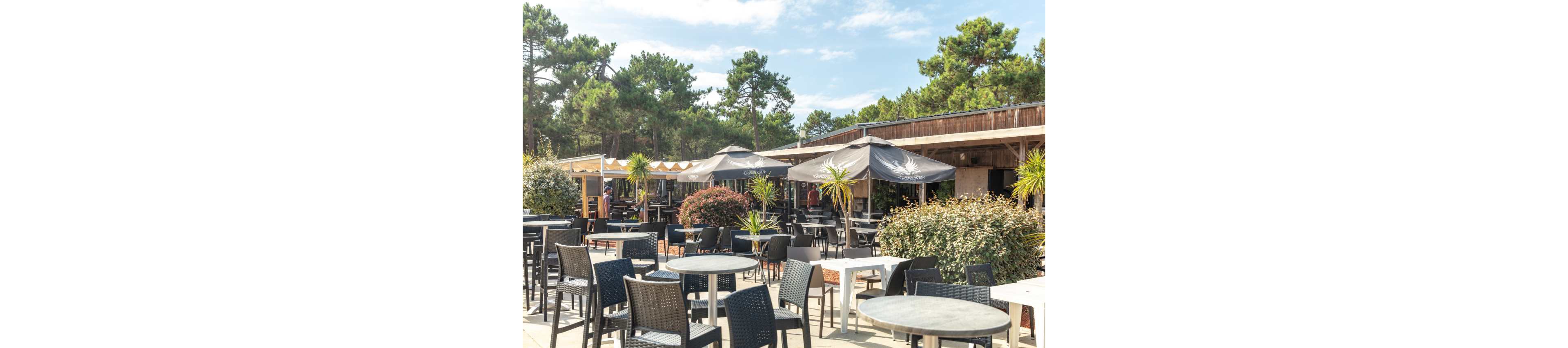 An outdoor café terrace features numerous empty tables and chairs arranged on a sunny patio. Large umbrellas provide shade near a wooden building surrounded by lush greenery and tall trees. A few people are visible in the background near the entrance area. The scene suggests a relaxed, vacation-like atmosphere in a natural setting.