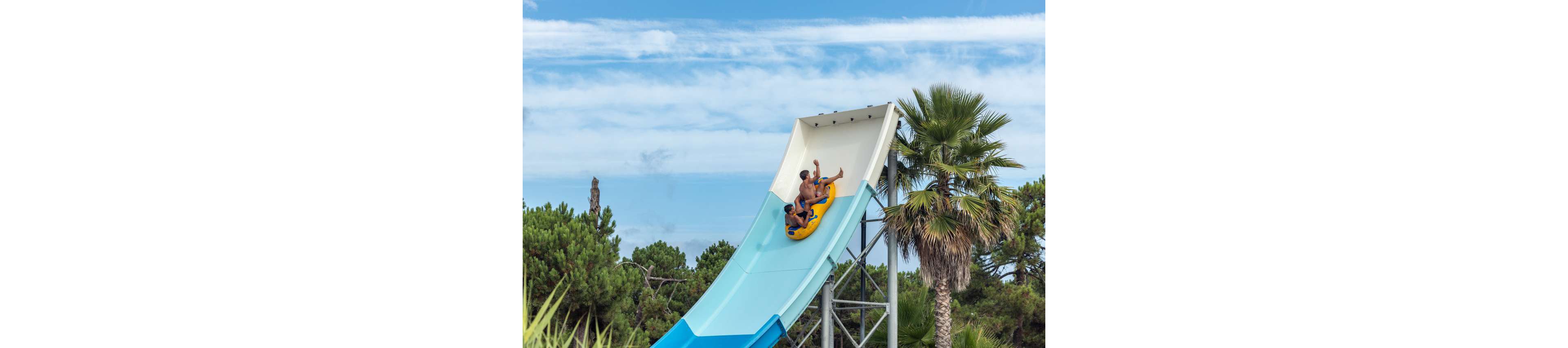 Two people ride together on a yellow inflatable raft down a tall blue waterslide. The slide structure rises above lush green trees and palm trees under a partly cloudy sky. One rider raises an arm in excitement as they descend. The scene captures a fun summer moment at an outdoor water park.