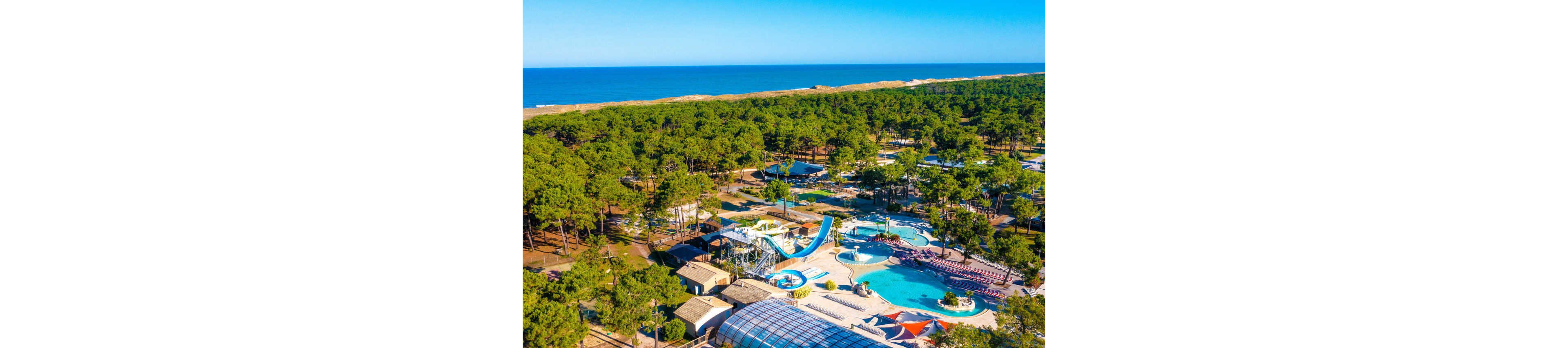 A large holiday resort with multiple outdoor swimming pools and water slides is nestled within a dense coastal pine forest. The aerial view shows curving pools, a tall slide structure, and sun loungers arranged around the water. A glass-roofed building and several low cabins or bungalows are visible among the trees. In the background, sand dunes and a calm blue sea stretch along the horizon under a clear sky.