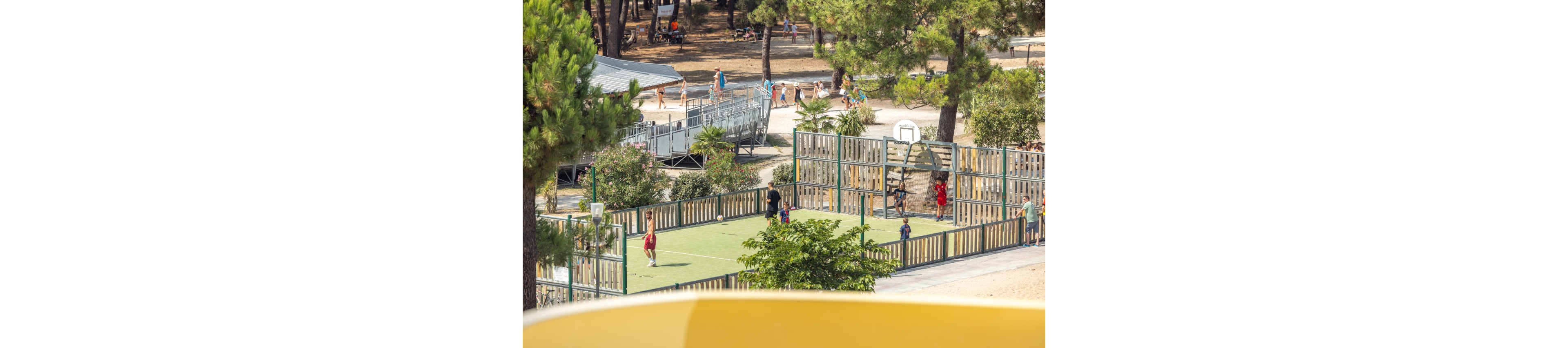 An outdoor multi-sport court is surrounded by wooden fencing and tall trees. Several children and adults are playing and walking around the court area. A basketball hoop is visible at one end, with people casually engaging in recreation. In the background, more visitors stroll along paths near tents or cabins in a sunny park or campground. The overall scene feels relaxed and family-friendly in a natural setting.