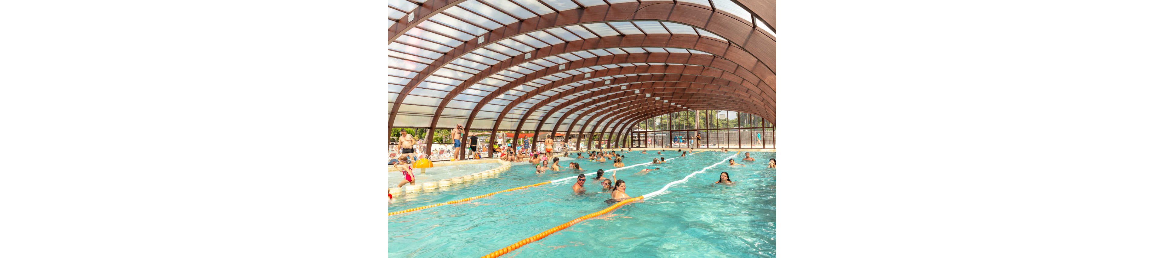 A large covered swimming pool is filled with people enjoying the water. Swimmers of various ages play, swim, and relax under an arched transparent roof. Sunlight filters through the structure, illuminating the turquoise pool and creating a bright, summery atmosphere. In the background, more visitors gather around the poolside area and outdoor surroundings.