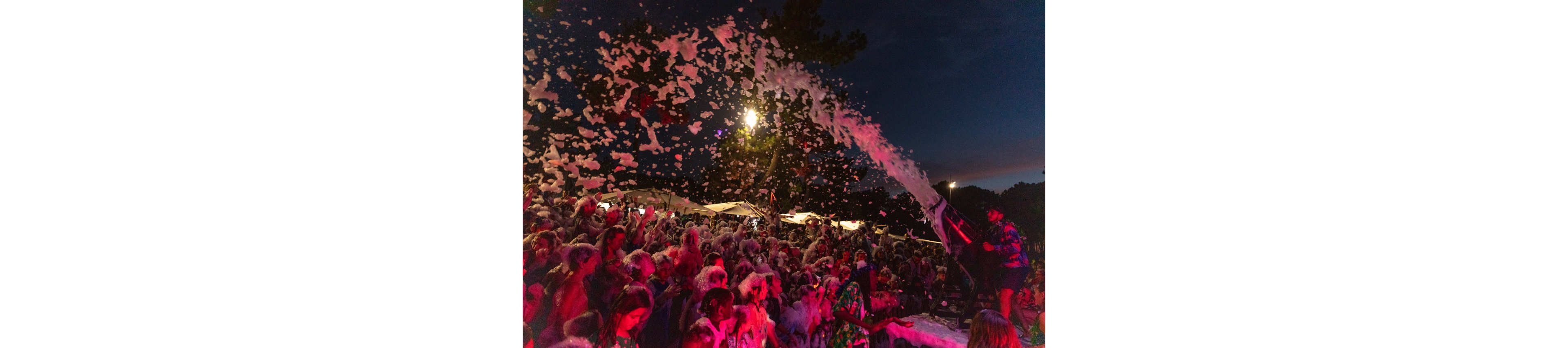 A large crowd of people enjoys a nighttime foam party outdoors. Thick streams of foam are sprayed over the audience from the stage, covering many participants. Colorful red and purple lights illuminate the scene against a dark evening sky. The atmosphere feels energetic, festive, and playful as people raise their hands and dance in the foam.