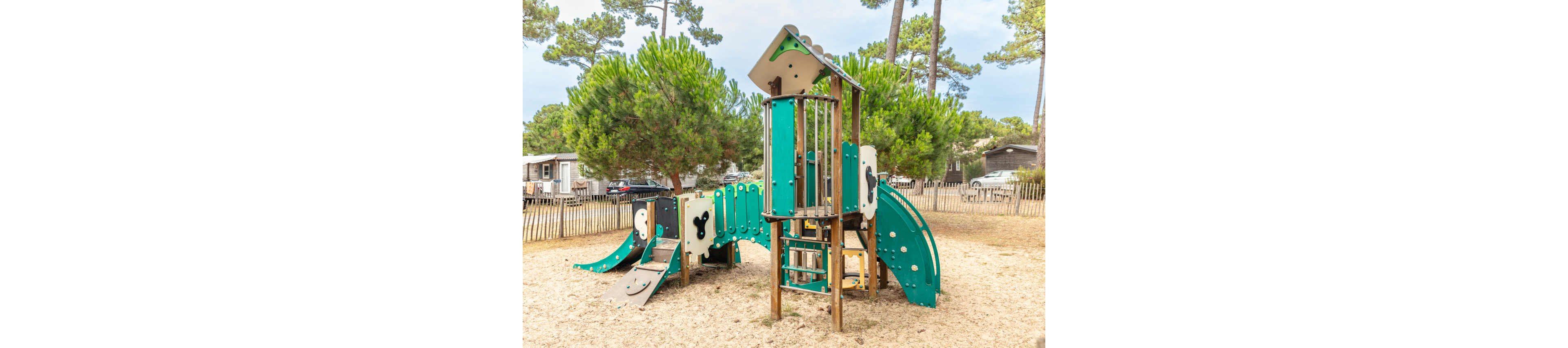 A modern green and beige playground structure stands on a sandy surface in an outdoor park. The play equipment includes slides, climbing panels, and a small tower with a ladder. Tall pine trees and wooden fences surround the play area, with parked cars and cabins visible in the background. The scene suggests a quiet, family-friendly recreational space in a natural setting.