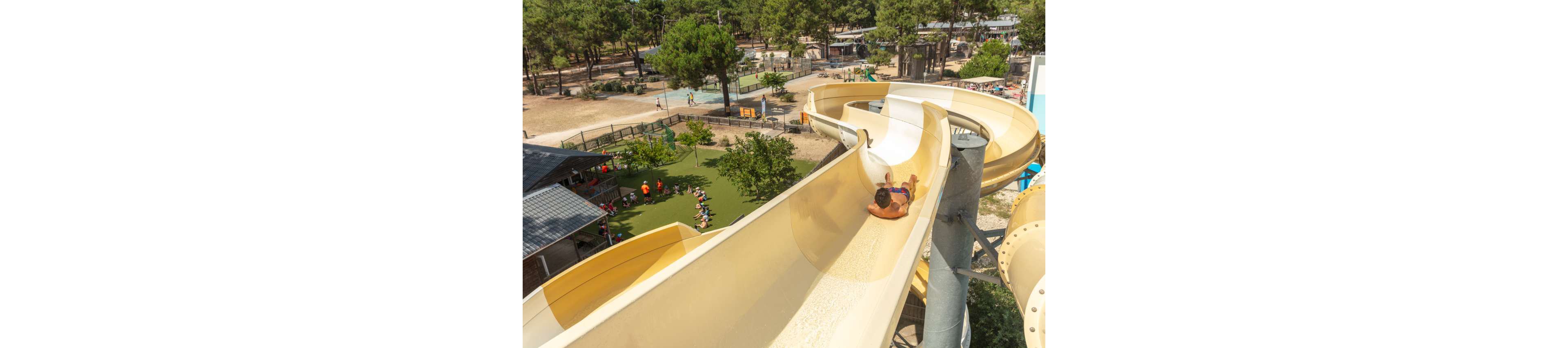 A person rides down a tall, winding waterslide at an outdoor water park. The slide curves above a landscaped area with grass, trees, and nearby buildings. Below, groups of people gather near shaded structures and recreational spaces. The scene is bright and sunny, suggesting a warm summer day at a family-friendly attraction.