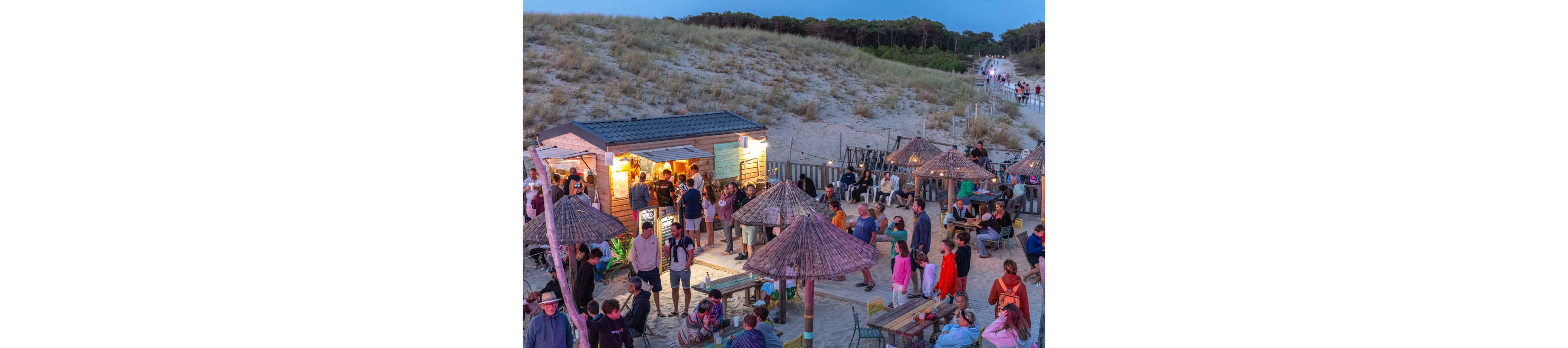 A lively crowd gathers around a small wooden beach bar nestled beside sand dunes at dusk. People sit at outdoor tables under straw parasols while others stand in line to order food and drinks. Warm string lights illuminate the bar area, contrasting with the cool evening sky. A sandy path and distant visitors lead toward the beach, creating a relaxed holiday atmosphere.