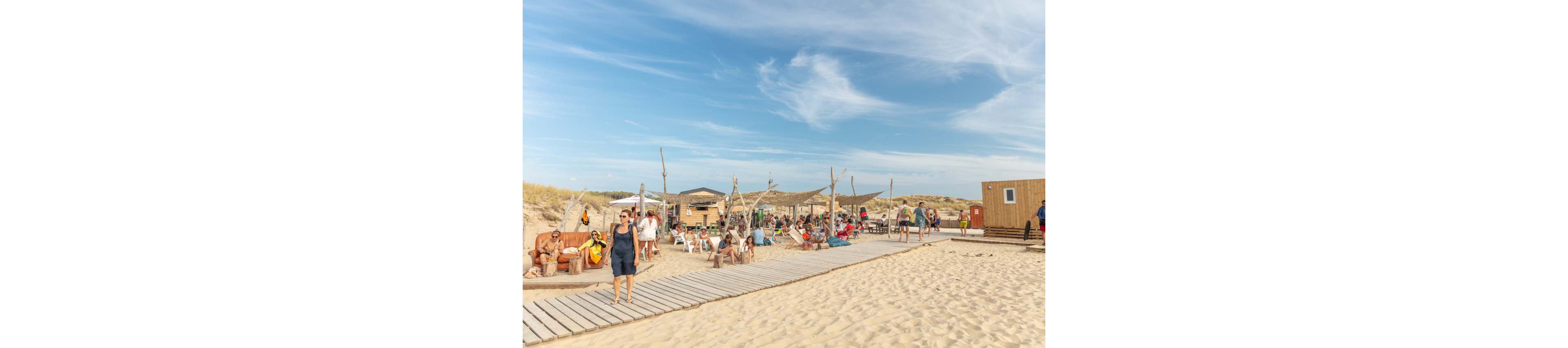 A group of people relax at a rustic beach bar set on a sandy shoreline. Wooden walkways and simple structures made of timber and canvas create a casual, bohemian atmosphere. Visitors sit on benches and chairs, chat, and enjoy the sunny weather under a bright blue sky with light clouds. Sand dunes and sparse vegetation frame the background, emphasizing the natural coastal setting.