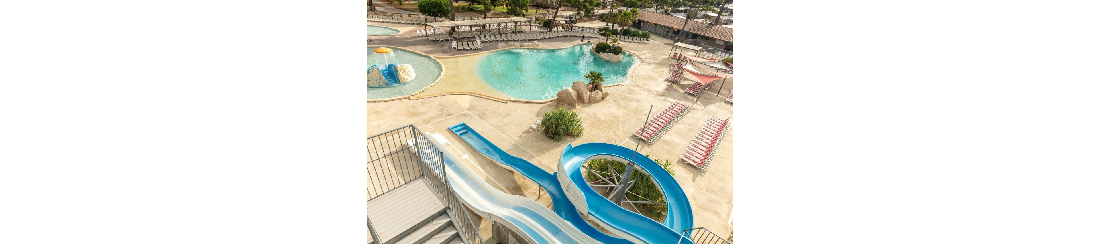 An elevated view overlooks a resort-style water park featuring a winding blue water slide leading into a pool. Curved swimming pools with shallow beach-style entries and rock formations create a relaxed vacation setting. Numerous empty lounge chairs and umbrellas surround the pools, suggesting an off-peak or early morning time. The overall scene feels sunny, calm, and inviting for families and holiday travelers.