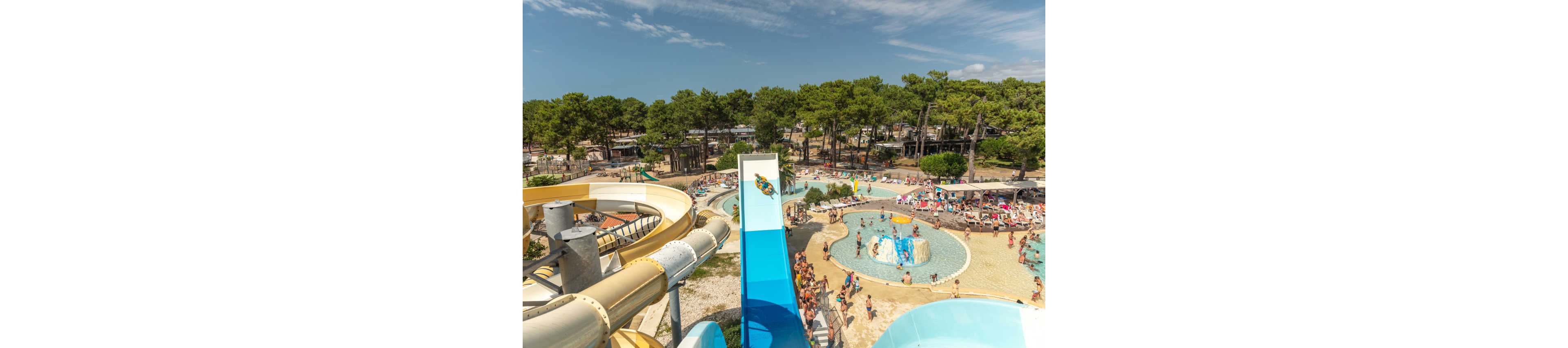 A large outdoor water park is filled with visitors enjoying multiple pools and winding water slides. People relax on sun loungers and under umbrellas while others line up for the tall slides. The scene is surrounded by dense green trees and clear blue sky, creating a holiday atmosphere. Bright colors from the slides, swimwear, and pool toys emphasize the fun, energetic summer mood.