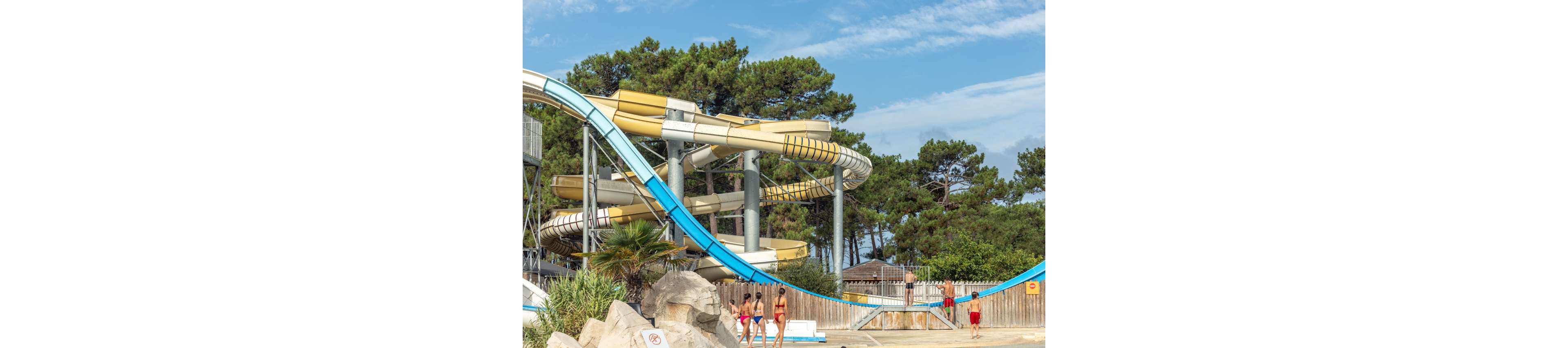 A group of people in swimwear walk near the base of large twisting water slides at an outdoor water park. The slides curve high above the ground, supported by metal pillars, with blue and beige tubes dominating the scene. Tall evergreen trees form a natural backdrop behind the park structures, and a wooden fence encloses the pool area. Bright daylight and clear skies create a summery, recreational atmosphere.