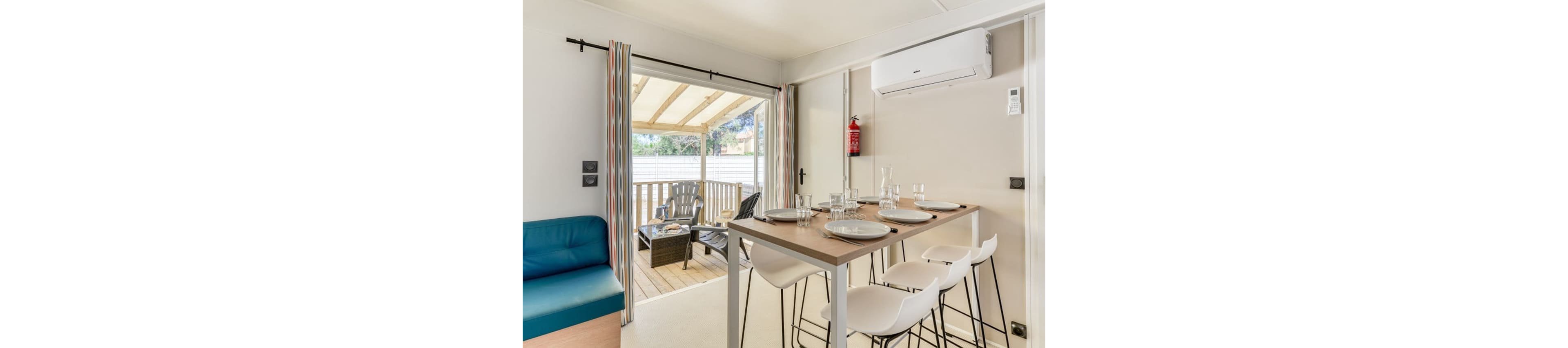 Compact modern dining area inside a light holiday cabin opens onto a covered wooden terrace. A tall table is set with plates, glasses, and cutlery beside white bar stools and a teal sofa bench. Outside, wicker chairs and a small table sit under a pergola-style roof with views of greenery. An air conditioning unit and a red fire extinguisher are mounted on the wall, emphasizing comfort and safety.
