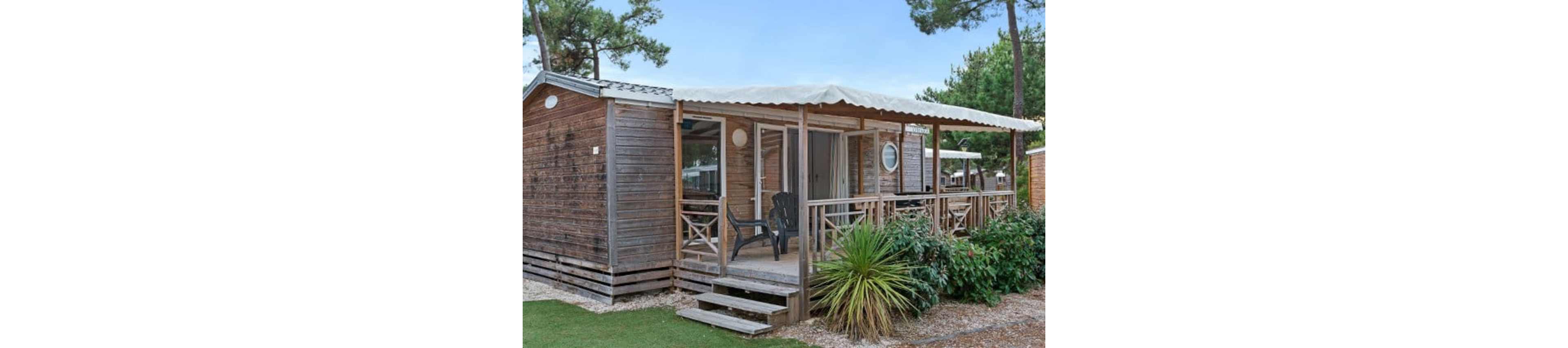 A small wooden cabin with a covered front terrace is shown in an outdoor setting. The structure features sliding glass doors, a few steps leading up to the deck, and plastic chairs arranged for seating. Surrounding the cabin are gravel paths, a patch of artificial grass, and various green plants. Tall trees and neighboring cabins in the background suggest a holiday park or campground environment.