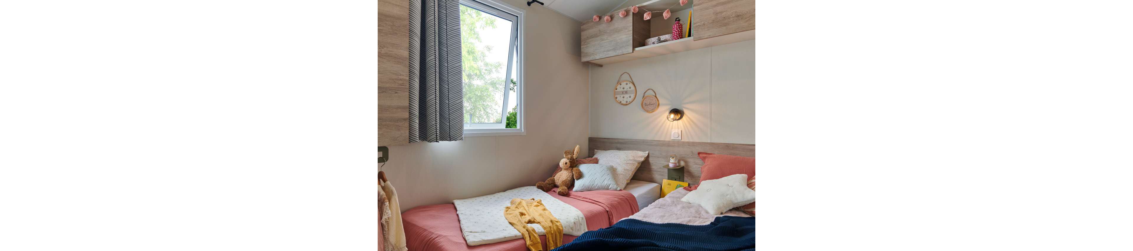 Small children's bedroom featuring two twin beds with soft pink and neutral bedding. A stuffed rabbit toy sits between the beds, adding a playful touch. Wall-mounted shelves and cabinets hold books and decorative items, while woven wall hangings and string lights create a warm atmosphere. Natural light enters through an open window with patterned curtains, brightening the compact, cozy space.
