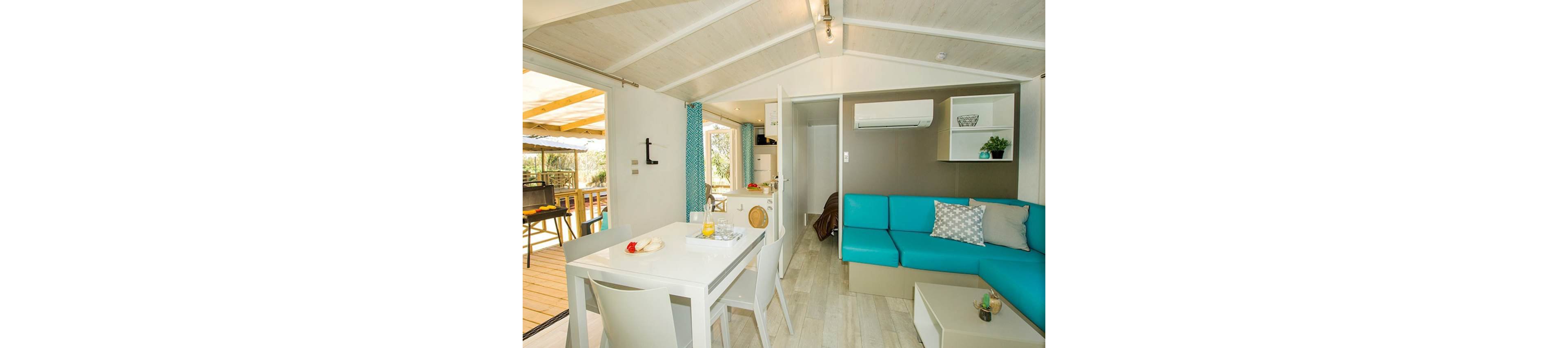 Compact open-plan living area in a modern holiday cabin with a turquoise corner sofa and white dining table. Large windows and a sliding door open to a wooden deck with outdoor seating and a grill. Light wood flooring and white paneled ceiling create an airy, coastal feel. An air conditioning unit, small shelves, and simple decor emphasize comfort and functionality for vacation living.