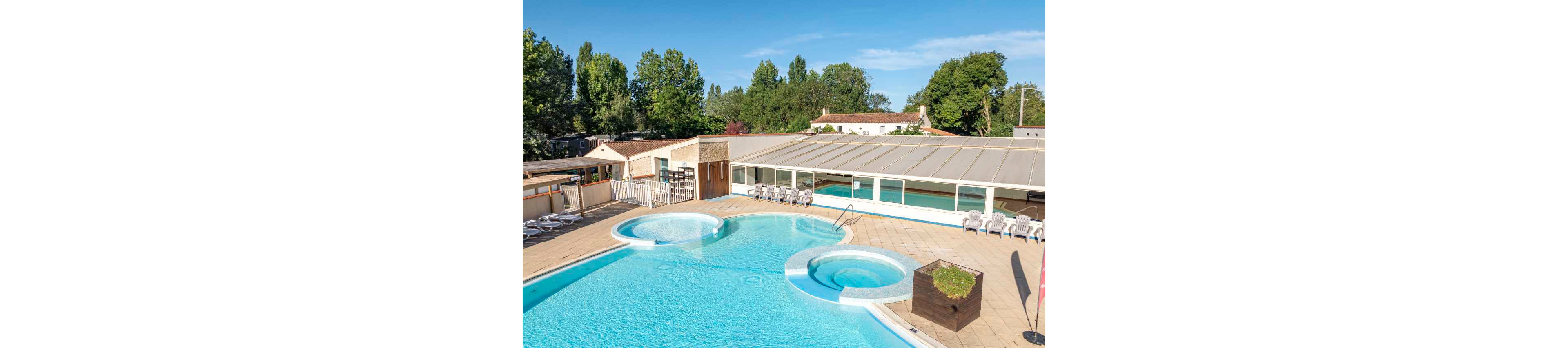 An outdoor swimming pool complex is shown on a bright sunny day at a holiday resort. The scene features a large main pool with curved edges and an attached circular wading pool. White lounge chairs line the pool deck, and a partially enclosed indoor pool area is visible behind large windows. Surrounding the facility are trees, low buildings, and clear blue sky, creating a relaxed vacation atmosphere.