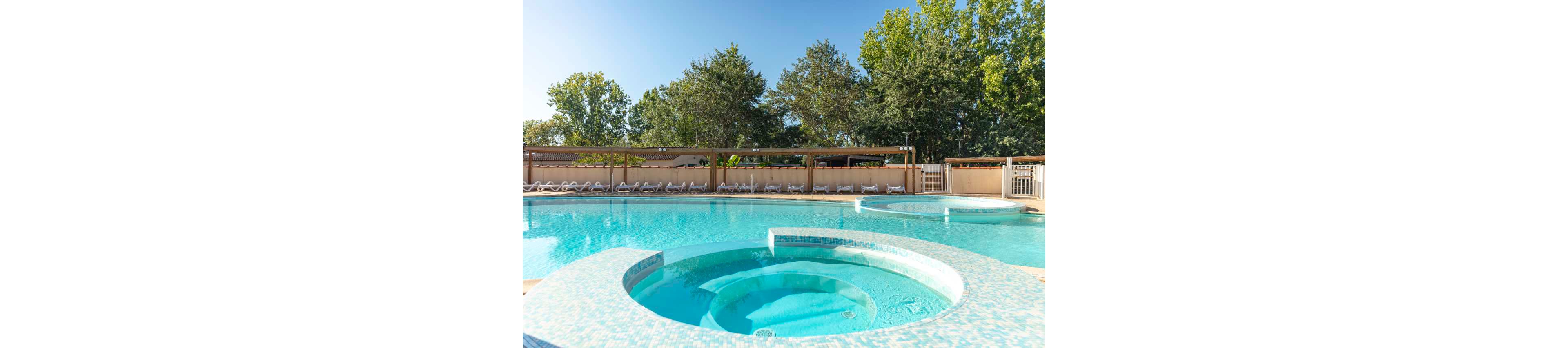 An outdoor resort-style swimming pool features a circular jacuzzi section in the foreground. The clear blue water contrasts with the light mosaic-tiled edges and curved pool shapes. In the background, rows of empty lounge chairs line the deck beneath a wooden pergola. Tall green trees and a bright blue sky create a sunny, relaxing vacation atmosphere.