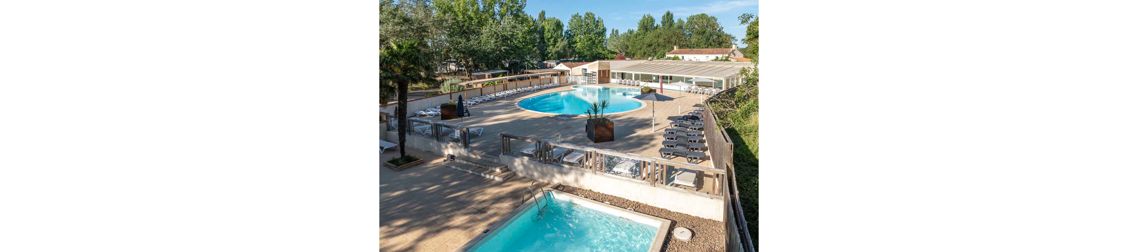 An outdoor resort pool area features a large circular swimming pool surrounded by numerous white sun loungers. A smaller rectangular pool sits on a lower level, separated by a fence and stone landscaping. The setting is bright and sunny, with clear blue water contrasting against the beige pool deck. Trees and low buildings in the background suggest a holiday park or campground environment.