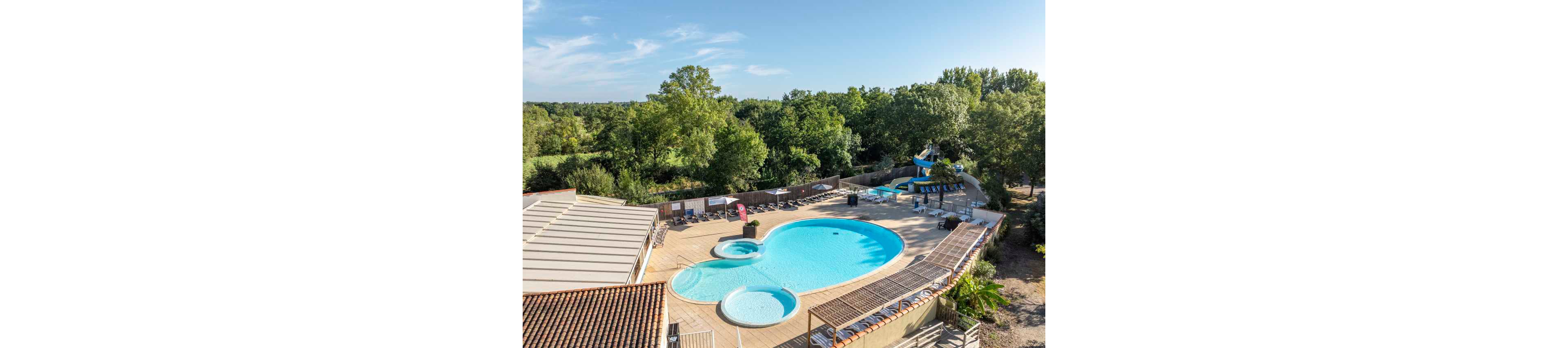 A large outdoor swimming pool complex is shown from an elevated viewpoint at a campsite or holiday park. The main heart-shaped pool is surrounded by sun loungers and parasols, with a smaller circular wading pool in the foreground. In the background, a curving blue waterslide ends in a separate pool area. The scene is framed by lush green trees and clear blue sky, suggesting a sunny summer day in a natural setting.
