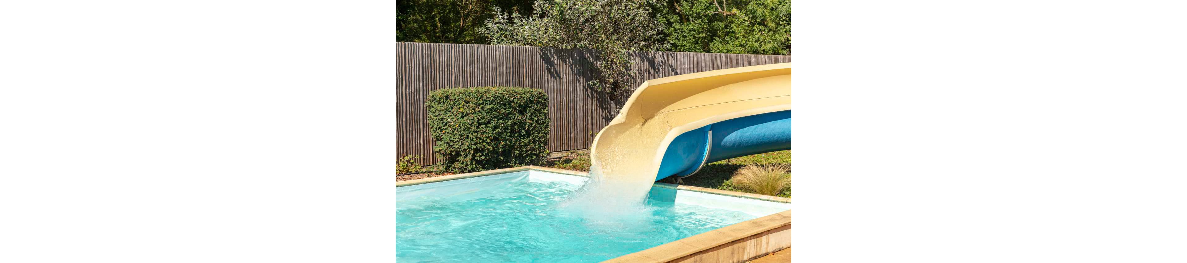 Two children enjoy a sunny day at an outdoor swimming pool with a large curved waterslide. One child is sliding down into the clear blue water, creating a splash, while another child waits in the pool. The scene is set in a fenced backyard area with green bushes and trees in the background. Bright daylight and colorful swimsuits emphasize a fun, playful summer atmosphere.