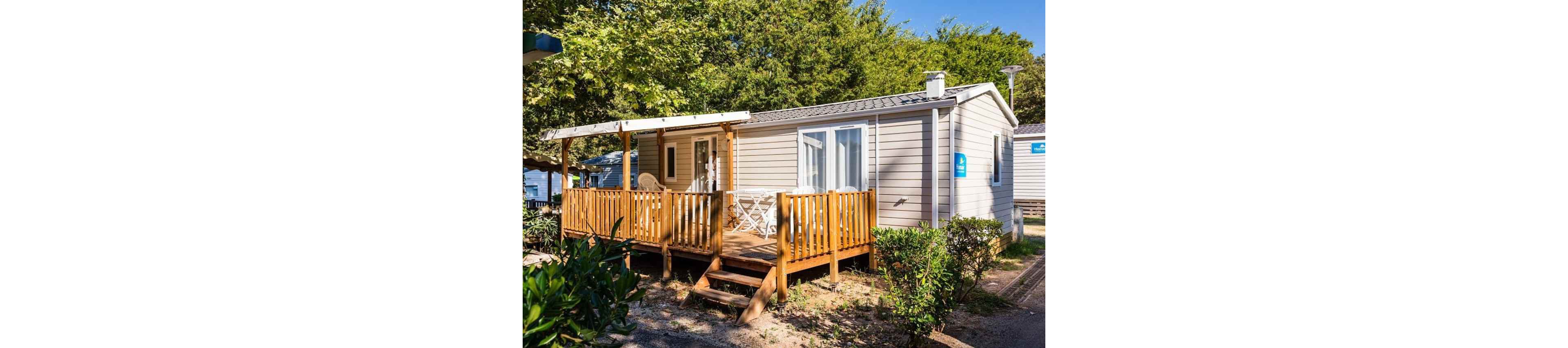 A light-colored mobile home features a small wooden deck with outdoor seating. The terrace is partially covered by a pergola, providing shade near the entrance door. White patio chairs and a table sit on the deck, suggesting a relaxed vacation setting. Lush green trees and shrubs surround the structure, creating a peaceful, natural atmosphere.