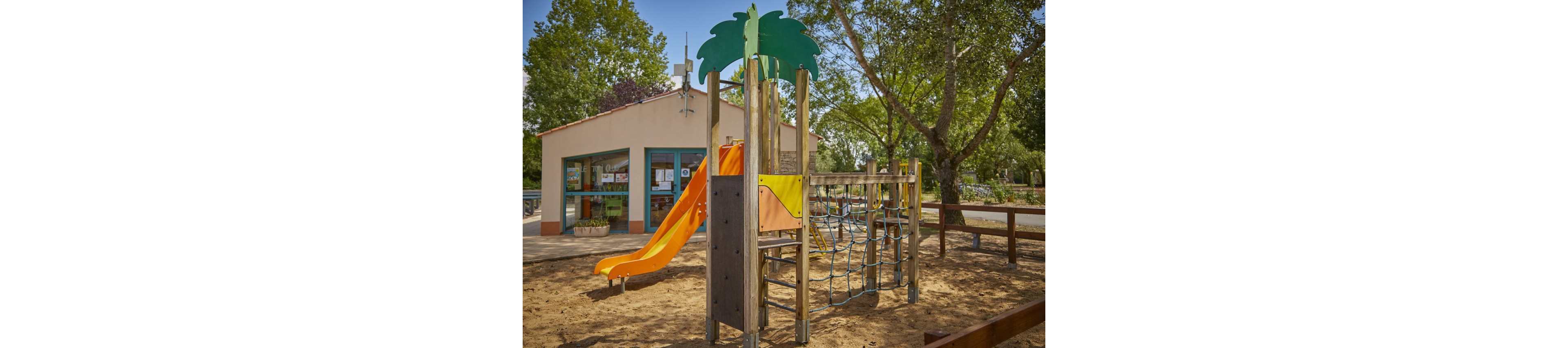 An outdoor playground features a bright orange slide and a wooden climbing structure with rope nets. The play area is set on sandy ground and surrounded by trees providing shade. In the background, a small building with large windows and colorful details suggests a park facility or community center. The scene is quiet and sunlit, evoking a relaxed family-friendly atmosphere.