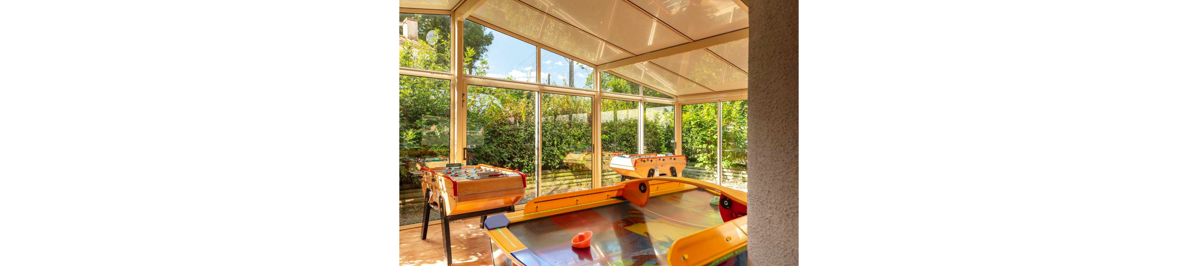 A bright indoor game room features colorful foosball tables and an air hockey table. Large floor-to-ceiling windows surround the space, revealing lush green trees and a garden outside. Warm sunlight fills the room, creating a cheerful and inviting atmosphere for play. The modern glass enclosure contrasts with the vibrant game tables and natural outdoor scenery.