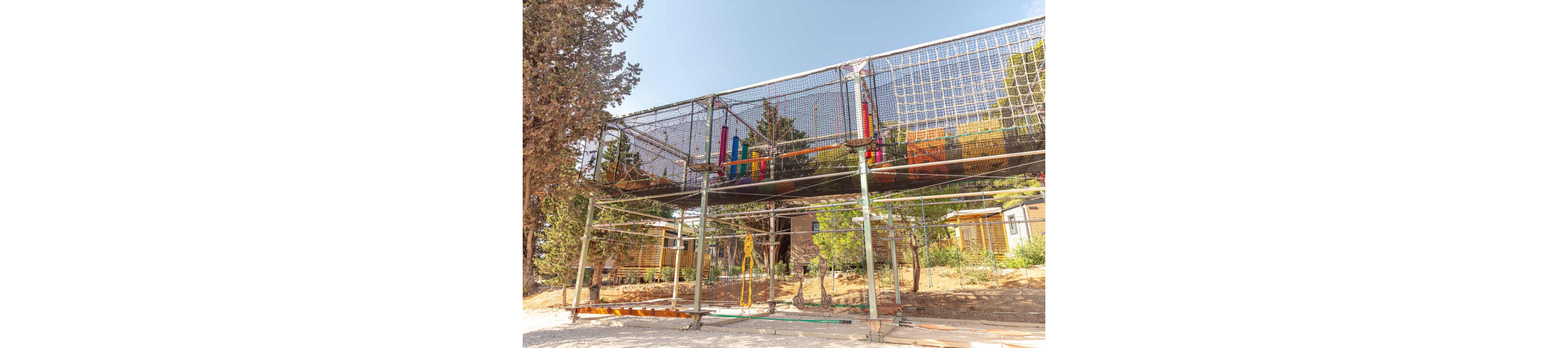 An elevated rope playground structure stands outdoors among tall trees. The play area is enclosed with safety netting and supported by metal scaffolding. Colorful padded obstacles and hanging elements are visible inside the structure. Wooden buildings and dry ground with sparse vegetation surround the playground under bright daylight.
