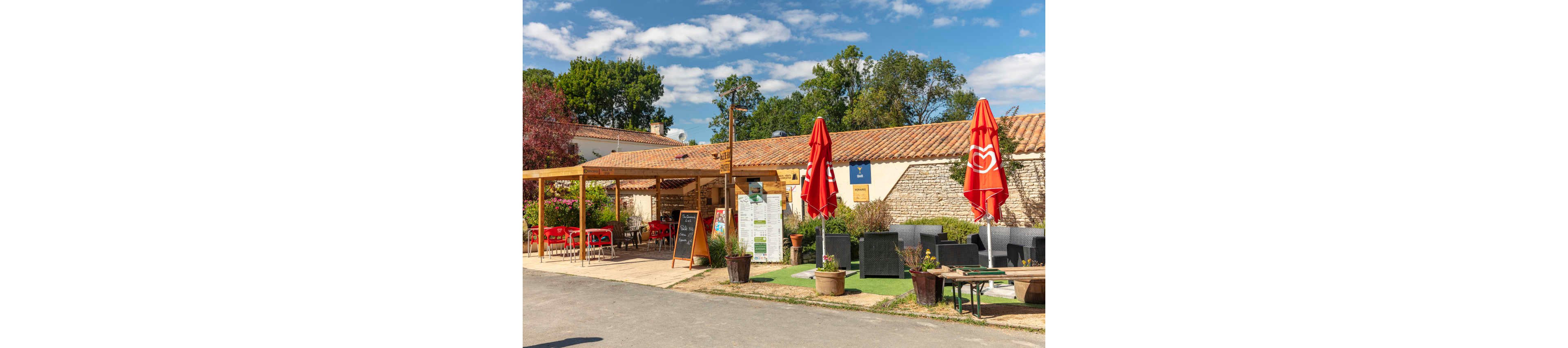 An outdoor café terrace is set against a rustic stone building with a tiled roof. Red umbrellas and matching chairs create a vibrant seating area under a wooden pergola. Potted plants, menu boards, and signs stand near the entrance, adding detail and inviting atmosphere. The scene is lit by bright daylight under a blue sky with scattered clouds, surrounded by green trees.